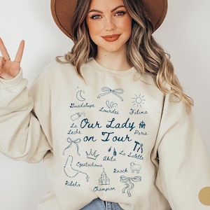 Puede incluir: Una sudadera de color crema con el texto "Our Lady on Tour" en letras azules. La sudadera presenta una variedad de iconos y lugares religiosos, incluyendo Guadalupe, Lourdes, F&aacute;tima, Par&iacute;s, Czestochowa, Akita, La Salette, Kibeho, Knock y Champion.