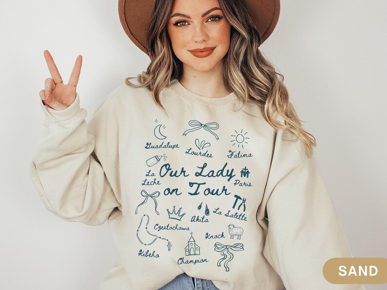 Puede incluir: Una sudadera de color crema con el texto "Our Lady on Tour" en letras azules. La sudadera presenta una variedad de iconos y lugares religiosos, incluyendo Guadalupe, Lourdes, F&aacute;tima, Par&iacute;s, Czestochowa, Akita, La Salette, Kibeho, Knock y Champion.