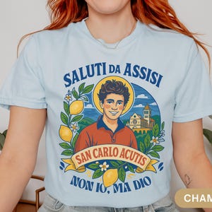 May include: Pale blue t-shirt with a graphic of San Carlo Acutis, lemons, and flowers, with Italian text. The shirt features the text "SALUTI DA ASSISI" and "NON IO, MA DIO".