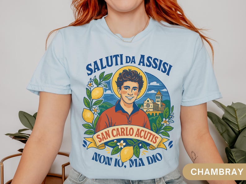 May include: Pale blue t-shirt with a graphic of San Carlo Acutis, lemons, and flowers, with Italian text. The shirt features the text "SALUTI DA ASSISI" and "NON IO, MA DIO".