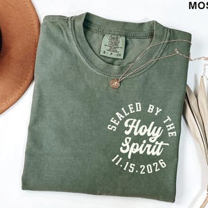 May include: Moss green Comfort Colors t-shirt with the text "SEALED BY THE Holy Spirit 11.15.2026" in a white circular design. A gold-colored necklace with a pendant is on the shirt. A brown hat is visible in the background.