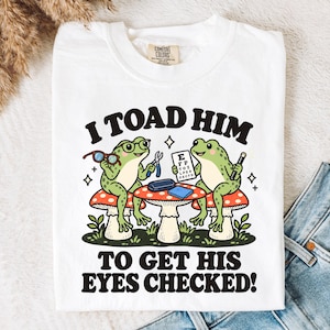 May include: A white t-shirt featuring a graphic of two green frogs sitting on mushroom stools. One frog wears glasses and holds a pair of spectacles, while the other holds an eye chart. The text reads I TOAD HIM TO GET HIS EYES CHECKED!