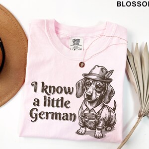 Puede incluir: Una camiseta de color rosa claro con un gr&aacute;fico marr&oacute;n oscuro de un perro salchicha con un sombrero tradicional y pantalones de cuero. El texto "I know a little German" est&aacute; impreso en una fuente marr&oacute;n oscuro a la izquierda de la ilustraci&oacute;n del perro. La camiseta tiene una etiqueta de la marca Comfort Colors.