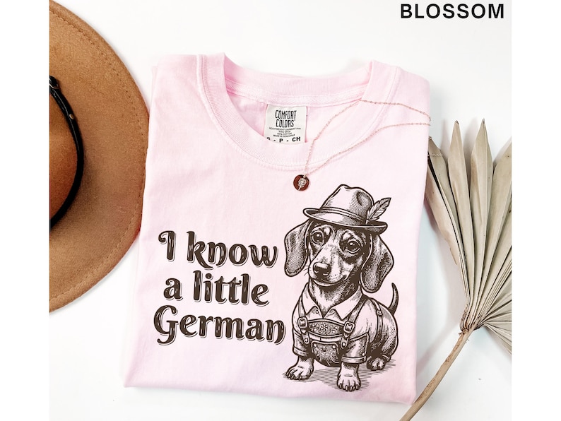Puede incluir: Una camiseta de color rosa claro con un gr&aacute;fico marr&oacute;n oscuro de un perro salchicha con un sombrero tradicional y pantalones de cuero. El texto "I know a little German" est&aacute; impreso en una fuente marr&oacute;n oscuro a la izquierda de la ilustraci&oacute;n del perro. La camiseta tiene una etiqueta de la marca Comfort Colors.