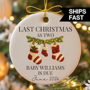 Op de afbeelding: Een witte keramische kerstversiering met de tekst "LAST CHRISTMAS AS TWO" en "BABY WILLIAMS IS DUE June 2026". De versiering heeft drie hangende kerstsokken in rood en crème kleuren, met een gouden lint om op te hangen.
