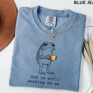 May include: A blue jean colored t-shirt featuring a cartoon frog holding a yellow mug. The frog has a weary expression. The text "God is still working on me" is printed below the frog. A gold necklace is draped across the shirt.