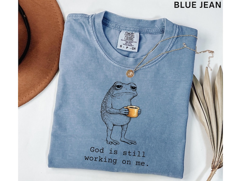 May include: A blue jean colored t-shirt featuring a cartoon frog holding a yellow mug. The frog has a weary expression. The text "God is still working on me" is printed below the frog. A gold necklace is draped across the shirt.