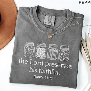 Puede incluir: Una camiseta color pimienta con un gr&aacute;fico blanco de cuatro frascos de vidrio llenos de conservas. Debajo de los frascos, el texto dice: "the Lord preserves his faithful. Psalm 31:23." La camiseta tiene cuello redondo y una etiqueta Comfort Colors.