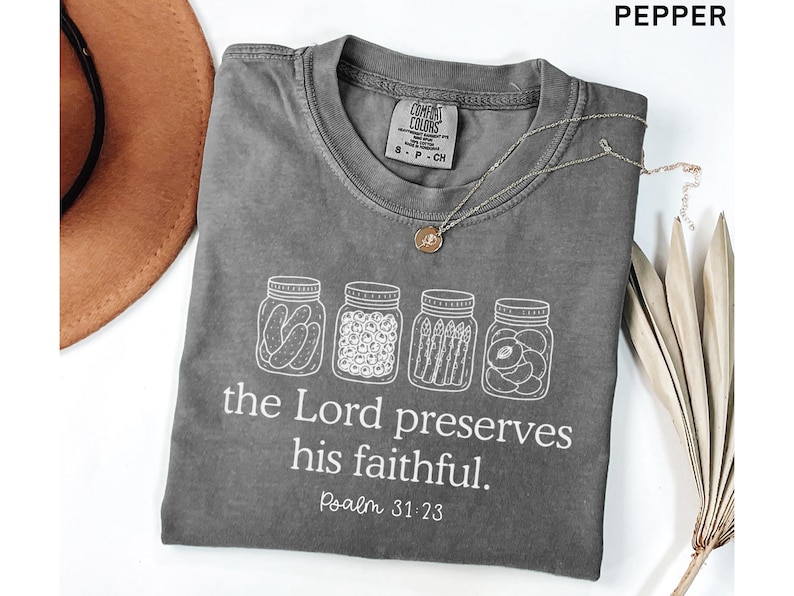 Puede incluir: Una camiseta color pimienta con un gr&aacute;fico blanco de cuatro frascos de vidrio llenos de conservas. Debajo de los frascos, el texto dice: "the Lord preserves his faithful. Psalm 31:23." La camiseta tiene cuello redondo y una etiqueta Comfort Colors.
