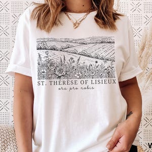 Pode incluir: Camiseta branca com uma ilustra&ccedil;&atilde;o em preto e branco de uma paisagem com campos, flores e o texto "ST. TH&Eacute;R&Egrave;SE OF LISIEUX ora pro nobis". A camiseta tem mangas curtas e uma bainha enrolada.