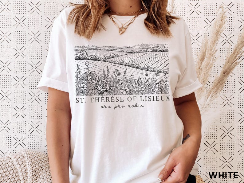 Pode incluir: Camiseta branca com uma ilustra&ccedil;&atilde;o em preto e branco de uma paisagem com campos, flores e o texto "ST. TH&Eacute;R&Egrave;SE OF LISIEUX ora pro nobis". A camiseta tem mangas curtas e uma bainha enrolada.