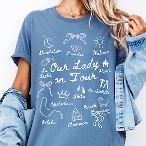 May include: A blue t-shirt with a white graphic design featuring the text "Our Lady on Tour" and various religious symbols and locations, including Guadalupe, Lourdes, Fatima, Paris, La Salette, Akita, Czestochowa, Kibeho, and Knock.