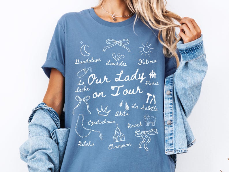 Puede incluir: Una camiseta azul con un dise&ntilde;o gr&aacute;fico blanco que presenta el texto "Our Lady on Tour" y varios s&iacute;mbolos y lugares religiosos, incluyendo Guadalupe, Lourdes, F&aacute;tima, Par&iacute;s, La Salette, Akita, Czestochowa, Kibeho y Knock.