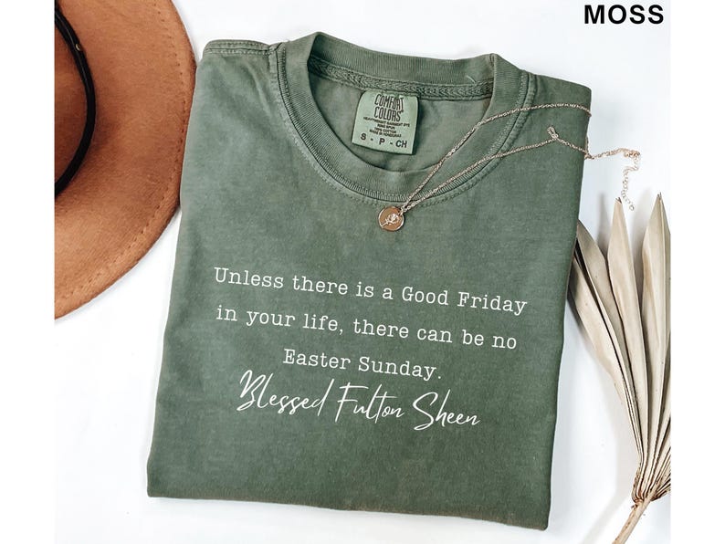 May include: A moss green t-shirt featuring the quote "Unless there is a Good Friday in your life, there can be no Easter Sunday. Blessed Fulton Sheen." A gold necklace and a brown hat are also visible.