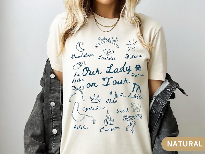 Puede incluir: Una camiseta beige con un dise&ntilde;o gr&aacute;fico azul que dice "Nuestra Se&ntilde;ora de gira" y presenta varios iconos y lugares religiosos, incluyendo Guadalupe, Lourdes, F&aacute;tima, Par&iacute;s, La Leche, Czestochowa, Akita, La Salette, Knock, Kibeho y Champion.