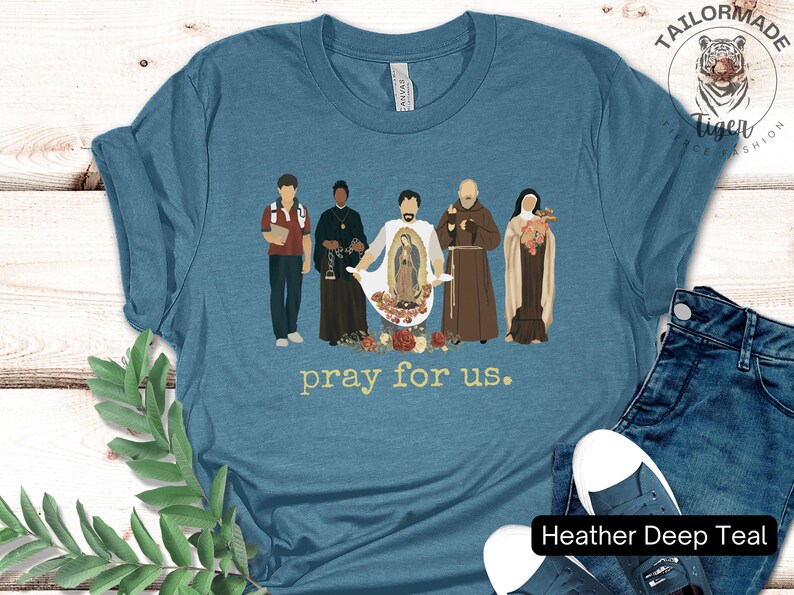 May include: A heather deep teal t-shirt with a graphic of five figures in front of a floral design. The figures are wearing robes and holding religious symbols. The text "pray for us." is printed below the graphic.