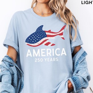 Puede incluir: Camiseta azul claro con un dise&ntilde;o de tibur&oacute;n con los colores de la bandera estadounidense. La palabra "AMERICA" est&aacute; impresa debajo del tibur&oacute;n, con "250 YEARS" debajo. La camiseta es de color azul claro.