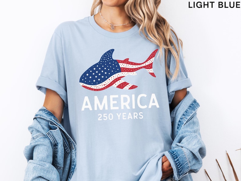 Puede incluir: Camiseta azul claro con un dise&ntilde;o de tibur&oacute;n con los colores de la bandera estadounidense. La palabra "AMERICA" est&aacute; impresa debajo del tibur&oacute;n, con "250 YEARS" debajo. La camiseta es de color azul claro.
