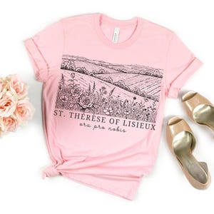 Pode incluir: Uma t-shirt rosa claro com uma ilustra&ccedil;&atilde;o a preto e branco de uma paisagem e o texto "ST. TH&Eacute;R&Egrave;SE OF LISIEUX ora pro nobis." A imagem inclui um ramo de rosas cor-de-rosa e sapatos de salto bege.