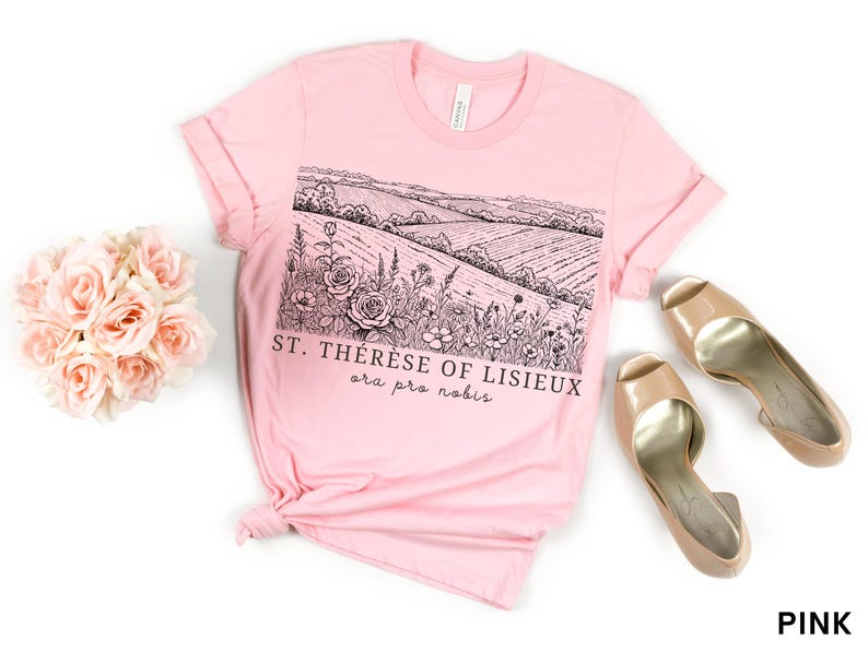 Pode incluir: Uma t-shirt rosa claro com uma ilustra&ccedil;&atilde;o a preto e branco de uma paisagem e o texto "ST. TH&Eacute;R&Egrave;SE OF LISIEUX ora pro nobis." A imagem inclui um ramo de rosas cor-de-rosa e sapatos de salto bege.