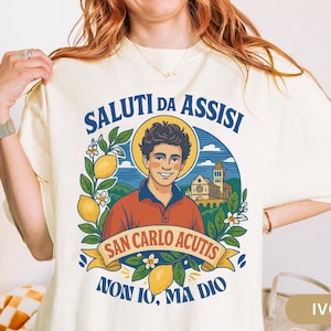 May include: Ivory t-shirt with a graphic of a smiling man with a halo, lemons, and the text "SALUTI DA ASSISI" and "SAN CARLO ACUTIS". The shirt also has the text "NON IO, MA DIO".