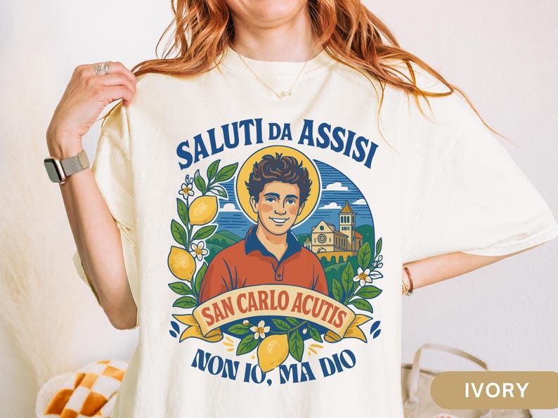 May include: Ivory t-shirt with a graphic of a smiling man with a halo, lemons, and the text "SALUTI DA ASSISI" and "SAN CARLO ACUTIS". The shirt also has the text "NON IO, MA DIO".