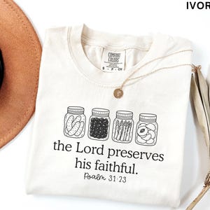 Puede incluir: Una camiseta de color marfil con un gr&aacute;fico de l&iacute;neas negras de cuatro frascos de vidrio llenos de pepinillos, bayas, esp&aacute;rragos y fruta en rodajas. Debajo de los frascos, el texto dice: the Lord preserves his faithful. Psalm 31:23.