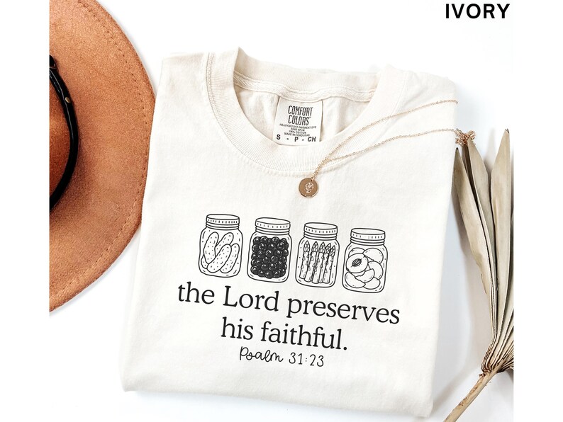 Puede incluir: Una camiseta de color marfil con un gr&aacute;fico de l&iacute;neas negras de cuatro frascos de vidrio llenos de pepinillos, bayas, esp&aacute;rragos y fruta en rodajas. Debajo de los frascos, el texto dice: the Lord preserves his faithful. Psalm 31:23.