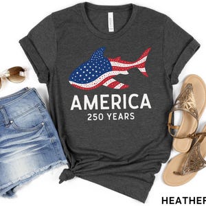Puede incluir: Camiseta gris oscuro con un dise&ntilde;o de tibur&oacute;n con los colores de la bandera estadounidense. La camiseta muestra la palabra "AMERICA" y "250 YEARS" en blanco. La imagen tambi&eacute;n incluye pantalones cortos vaqueros, gafas de sol y sandalias.