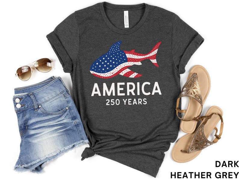 Puede incluir: Camiseta gris oscuro con un dise&ntilde;o de tibur&oacute;n con los colores de la bandera estadounidense. La camiseta muestra la palabra "AMERICA" y "250 YEARS" en blanco. La imagen tambi&eacute;n incluye pantalones cortos vaqueros, gafas de sol y sandalias.
