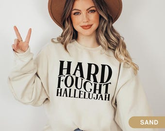 May include: Sand-colored sweatshirt with the words "HARD FOUGHT HALLELUJAH" in bold black letters. The model is wearing a brown hat and making a peace sign with their fingers.