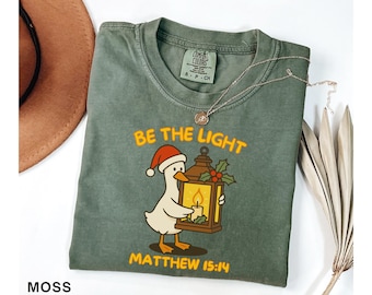 May include: Moss green t-shirt with a graphic of a duck wearing a Santa hat and holding a lantern with a lit candle. Text reads "Be the Light" and "Matthew 15:14". A brown hat and necklace are also visible.