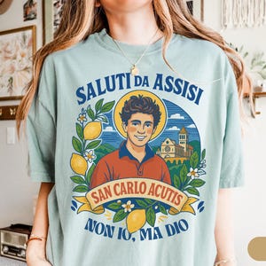 May include: A pale blue t-shirt with a graphic of a smiling man with a halo, lemons, and flowers, and the text "SALUTI DA ASSISI" and "SAN CARLO ACUTIS". The shirt has a red collar.