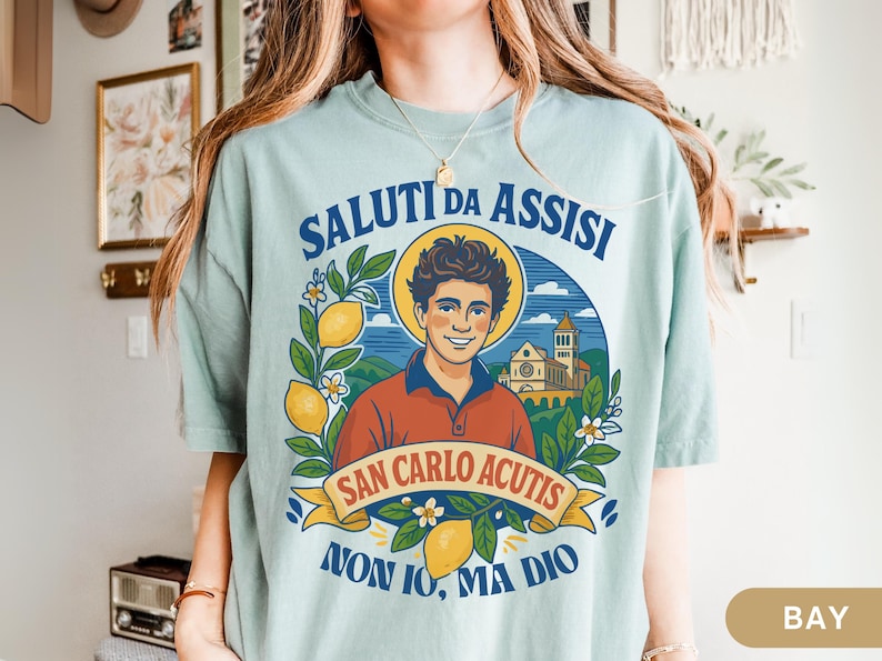 May include: A pale blue t-shirt with a graphic of a smiling man with a halo, lemons, and flowers, and the text "SALUTI DA ASSISI" and "SAN CARLO ACUTIS". The shirt has a red collar.