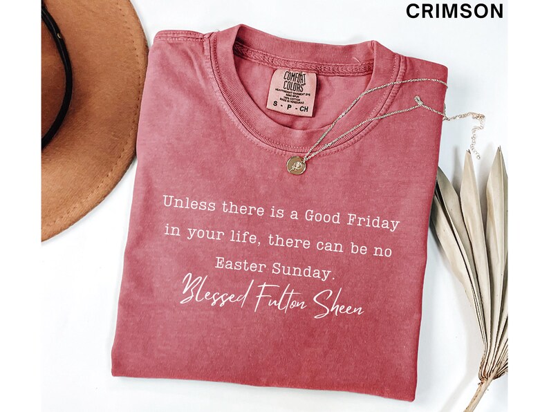 May include: Crimson t-shirt featuring the quote "Unless there is a Good Friday in your life, there can be no Easter Sunday. Blessed Fulton Sheen." A gold pendant necklace and a brown hat are also visible.