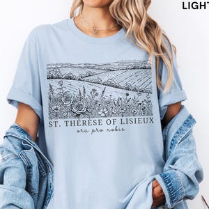 Pode incluir: Camiseta azul clara com uma ilustra&ccedil;&atilde;o a preto e branco de uma paisagem de campo com flores e o texto "ST. TH&Eacute;R&Egrave;SE OF LISIEUX ora pro nobis". A camiseta tem mangas curtas e um corte folgado.