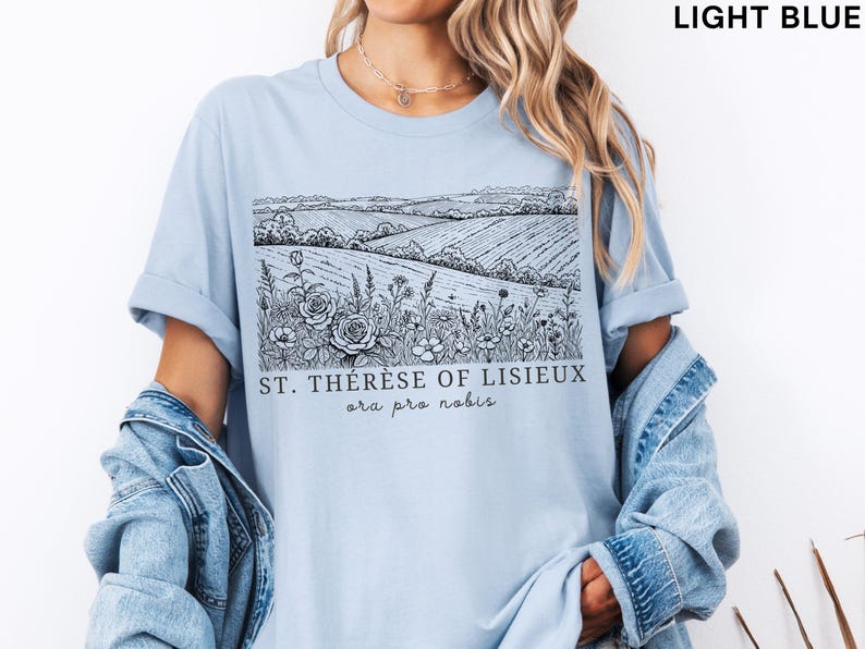 Pode incluir: Camiseta azul clara com uma ilustra&ccedil;&atilde;o a preto e branco de uma paisagem de campo com flores e o texto "ST. TH&Eacute;R&Egrave;SE OF LISIEUX ora pro nobis". A camiseta tem mangas curtas e um corte folgado.