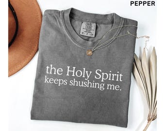 Könnte beinhalten: Ein anthrazitfarbenes Kurzarm-T-Shirt mit Rundhalsausschnitt und dem weißen Text "the Holy Spirit keeps shushing me." auf der Vorderseite. Das Shirt hat ein Comfort Colors Markenetikett am Innenkragen sowie eine goldfarbene Anhängerkette.