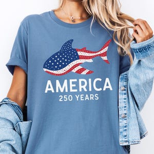 Puede incluir: Camiseta azul acero con un dise&ntilde;o de tibur&oacute;n con el patr&oacute;n de la bandera estadounidense. La palabra "AMERICA" est&aacute; impresa en blanco, con "250 YEARS" debajo. La camiseta tiene un corte relajado.
