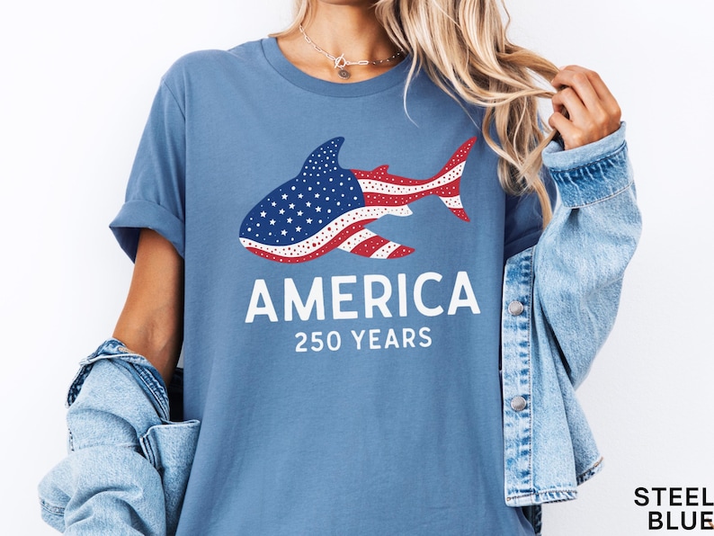 Puede incluir: Camiseta azul acero con un dise&ntilde;o de tibur&oacute;n con el patr&oacute;n de la bandera estadounidense. La palabra "AMERICA" est&aacute; impresa en blanco, con "250 YEARS" debajo. La camiseta tiene un corte relajado.