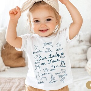 Puede incluir: Camiseta blanca para beb&eacute; con texto azul e ilustraciones. El texto dice "Nuestra Se&ntilde;ora en gira" e incluye nombres de varios sitios de peregrinaci&oacute;n cat&oacute;lica, incluyendo Guadalupe, Lourdes, F&aacute;tima, Par&iacute;s, La Leche, Akita, La Salette, Czutochows, Knock, Kileho y Champion.