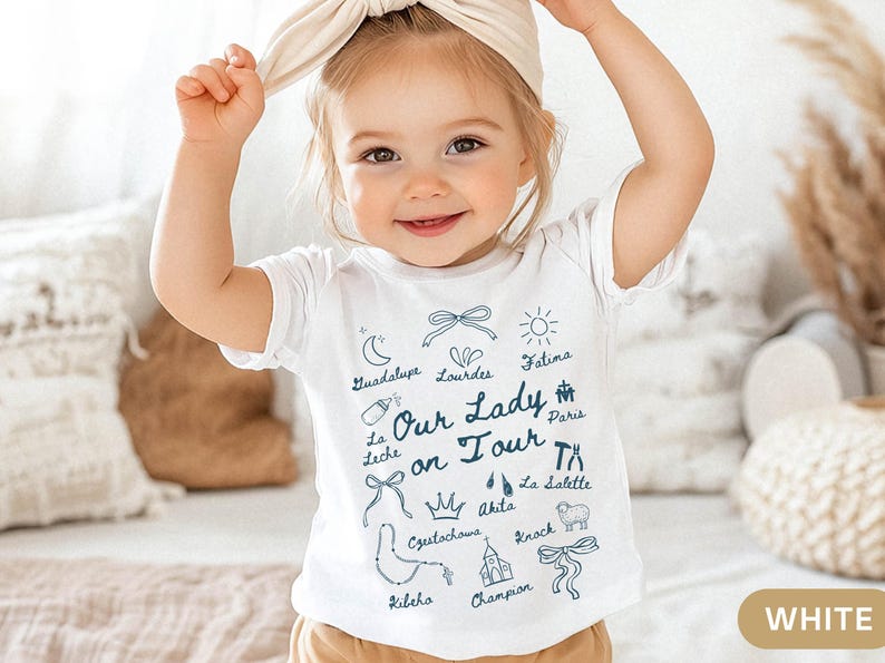 Puede incluir: Camiseta blanca para beb&eacute; con texto azul e ilustraciones. El texto dice "Nuestra Se&ntilde;ora en gira" e incluye nombres de varios sitios de peregrinaci&oacute;n cat&oacute;lica, incluyendo Guadalupe, Lourdes, F&aacute;tima, Par&iacute;s, La Leche, Akita, La Salette, Czutochows, Knock, Kileho y Champion.