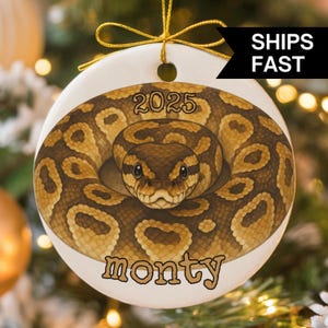 Op de afbeelding: Witte keramische kerstversiering met een illustratie van een opgerolde slang. De slang is bruin en bruin met een gedetailleerd patroon. De versiering heeft de tekst "2025" en "monty" in een decoratief lettertype. De tekst "SHIPS FAST" is ook zichtbaar.