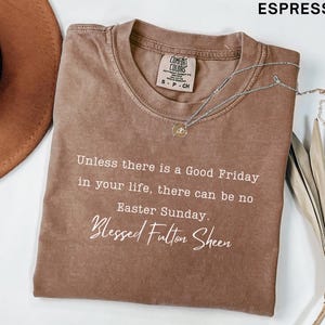 May include: Espresso-coloured t-shirt with the text "Unless there is a Good Friday in your life, there can be no Easter Sunday. Blessed Fulton Sheen." A silver necklace with a pendant rests on the shirt. A brown hat is visible.