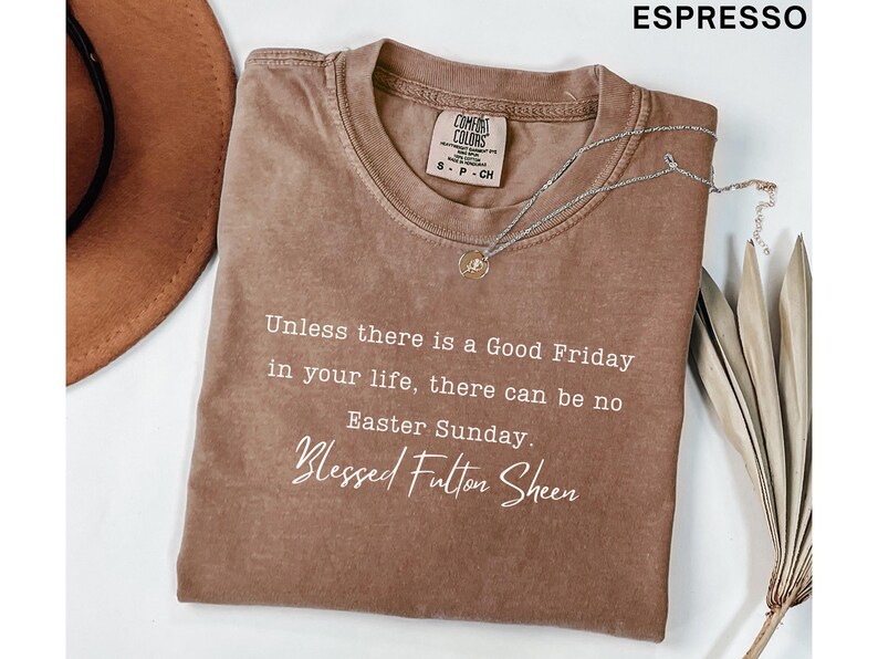 May include: Espresso-coloured t-shirt with the text "Unless there is a Good Friday in your life, there can be no Easter Sunday. Blessed Fulton Sheen." A silver necklace with a pendant rests on the shirt. A brown hat is visible.