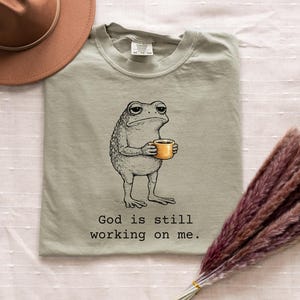 May include: Sage green t-shirt featuring a cartoon frog holding a coffee cup. The frog has a grumpy expression. The text "God is still working on me" is printed below the frog in black. A brown hat and dried flowers are also in the image.