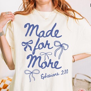 Puede incluir: Camiseta de color marfil con la frase "Made for More" en azul y dibujos de lazos. El texto "Efesios 2:10" tambi&eacute;n est&aacute; impreso en la camiseta. La camiseta es de estilo casual y c&oacute;modo.