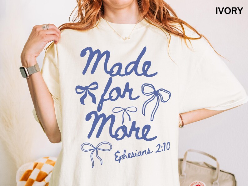 Puede incluir: Camiseta de color marfil con la frase "Made for More" en azul y dibujos de lazos. El texto "Efesios 2:10" tambi&eacute;n est&aacute; impreso en la camiseta. La camiseta es de estilo casual y c&oacute;modo.