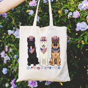 以下が含まれることがあります： 3匹のジャーマンシェパードが描かれた白いトートバッグ。各犬は、赤、白、青の愛国的な帽子とバンダナを着用しています。星とストライプがバッグの底を飾り、アメリカの誇りを祝っています。