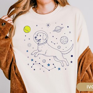 May include: A white sweatshirt with a graphic of a dog wearing a space helmet and floating in space. The graphic also includes stars, planets, and a tennis ball. The text "IVORY" is visible in the lower right corner of the image.